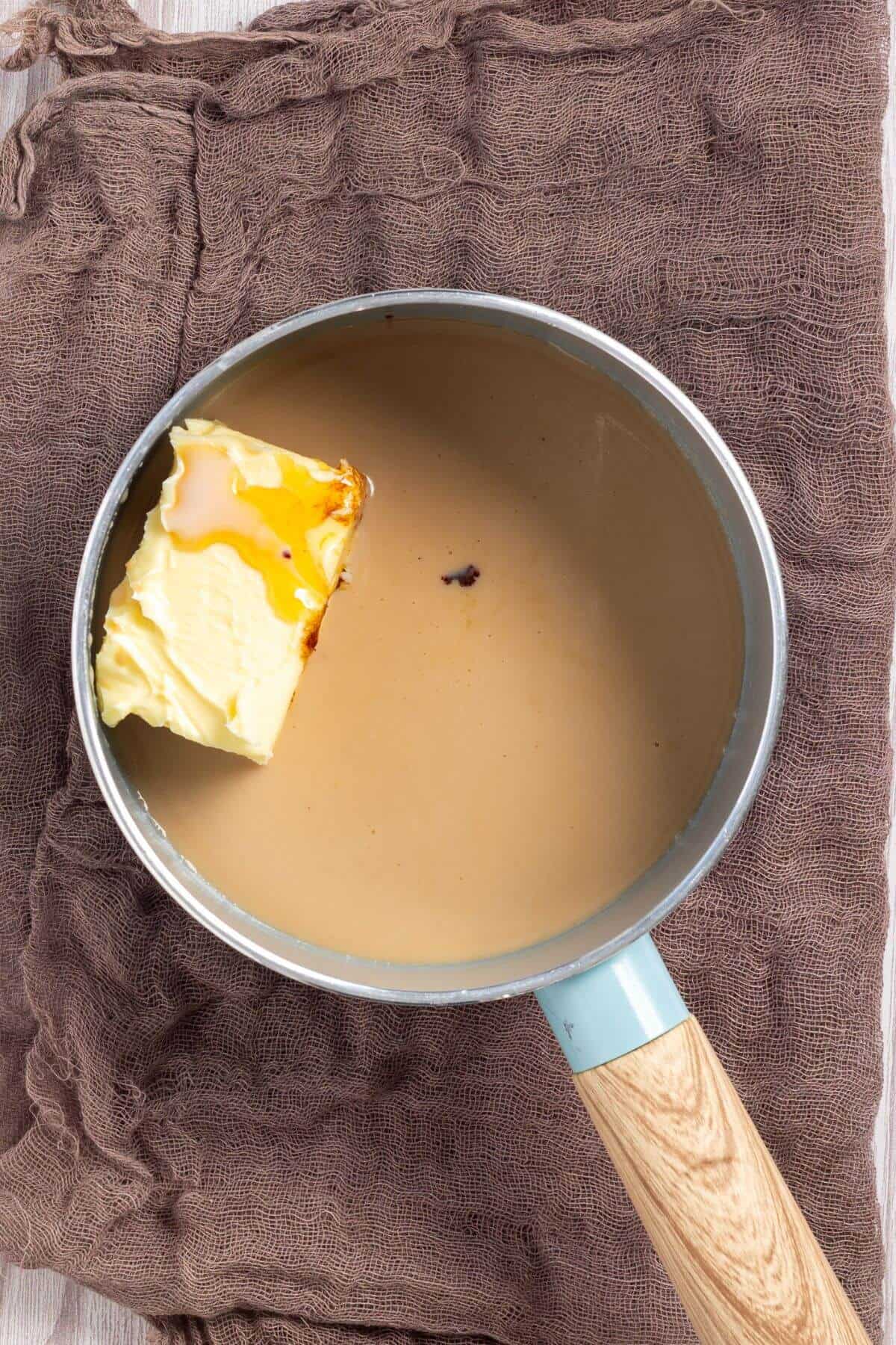 A saucepot contains a light brown liquid and a block of butter on a wooden table surface.