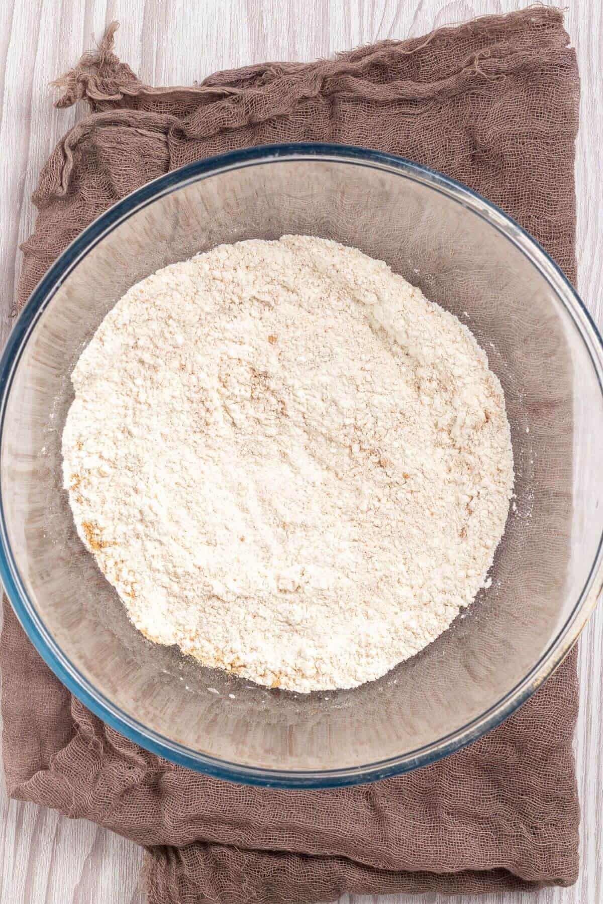 A glass bowl filled with flour rests on a brown cloth on a wooden surface.