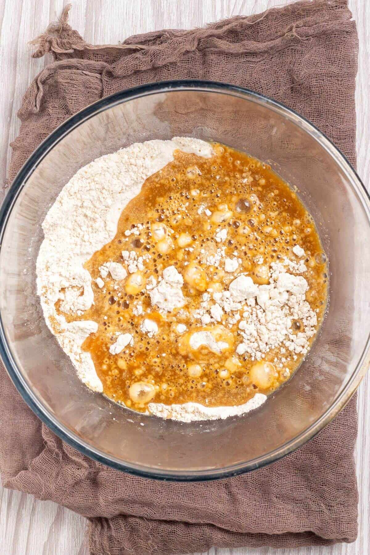 A glass bowl containing flour and liquid ingredients on a brown cloth.