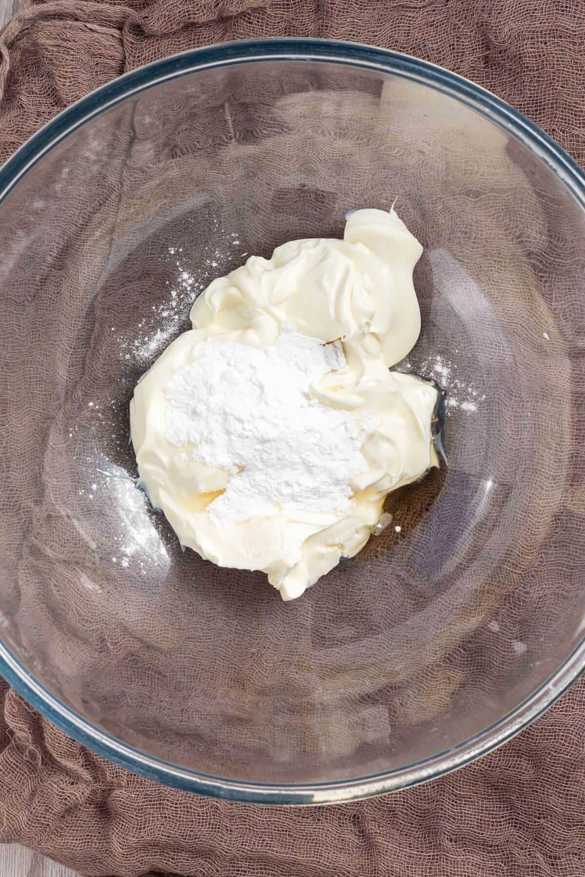 A glass bowl containing cream cheese and powdered sugar on a brown cloth.