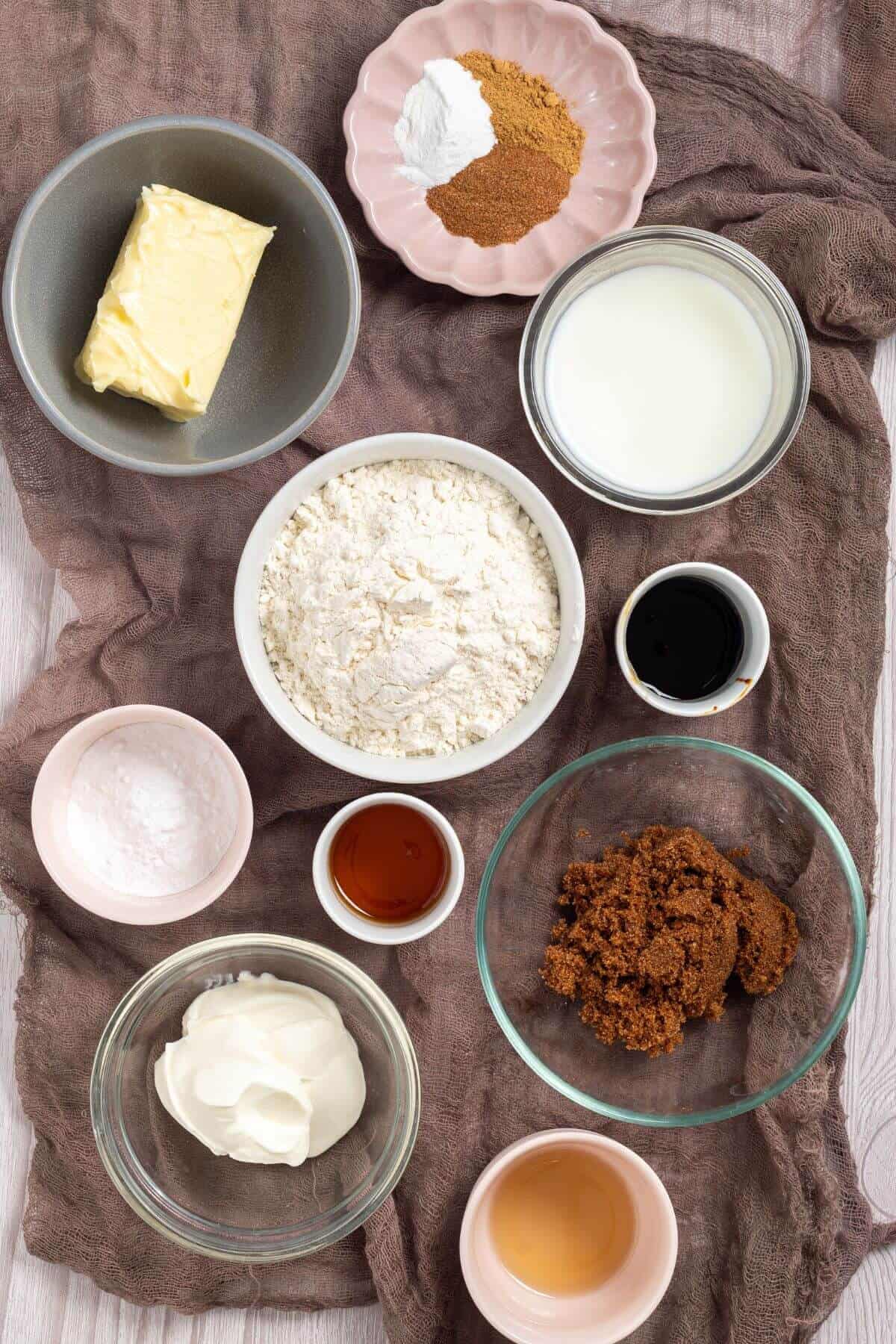A bird's-eye view of baking ingredients on a cloth: butter, flour, milk, spices, brown sugar, vanilla, yogurt, baking soda, and vinegar arranged in bowls on a wooden surface.