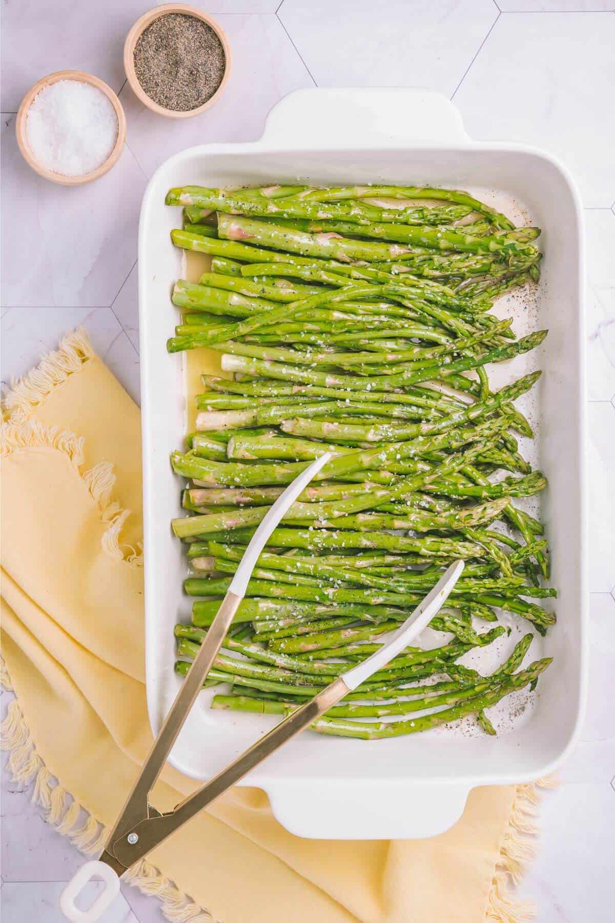 A white baking dish with roasted asparagus spears, garnished with salt, next to a pair of tongs. Small bowls of salt and pepper are in the background, with a yellow cloth underneath.