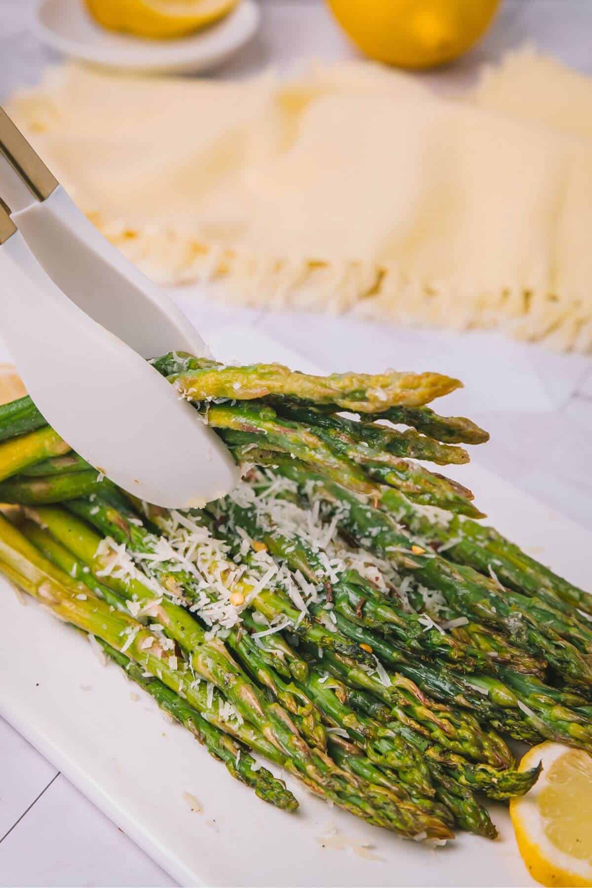 Tongs holding roasted asparagus topped with grated cheese on a white plate, with lemon slices nearby.