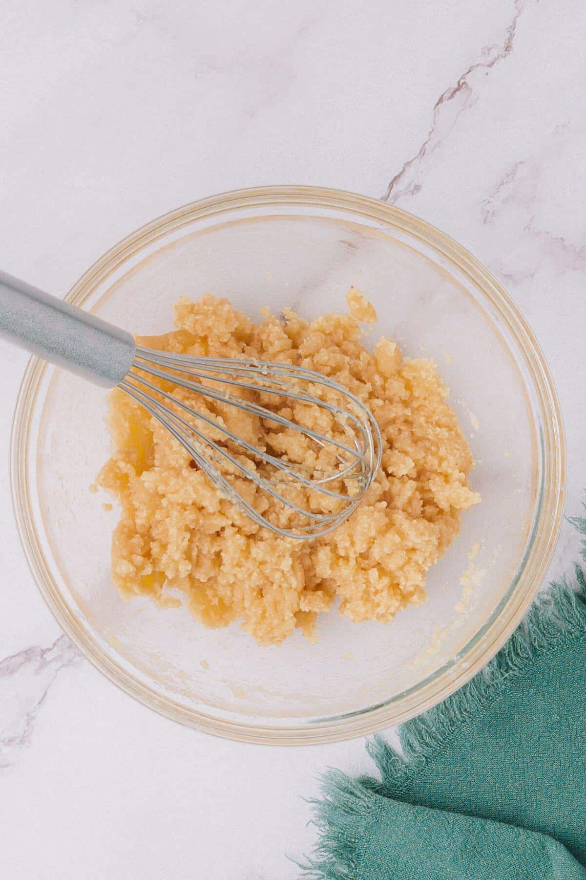 A glass bowl with a whisk and a mixture of wet ingredients on a marble countertop, next to a green cloth.