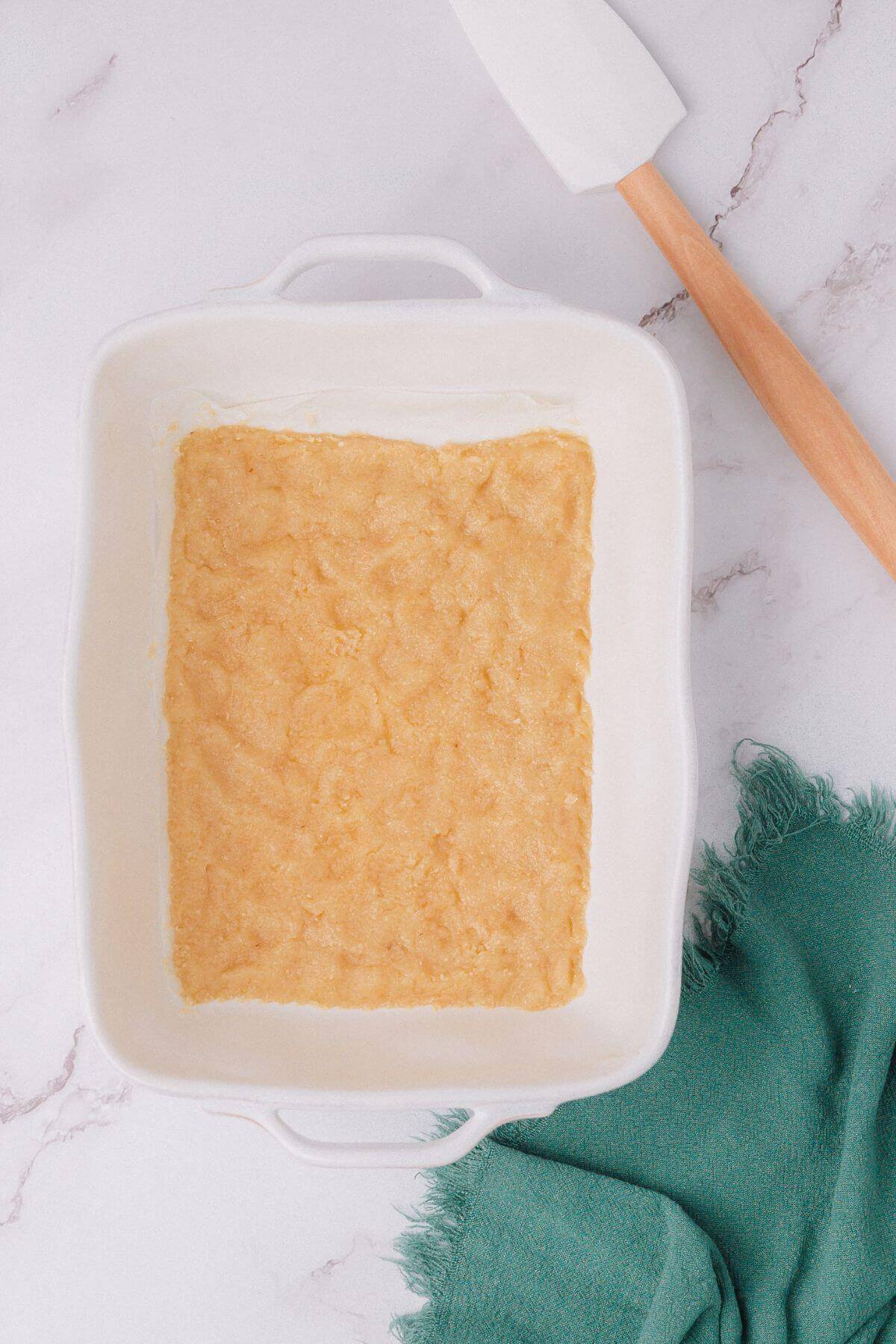 A rectangular white baking dish with a layer of dough is on a marble surface, next to a white spatula with a wooden handle and a green cloth.