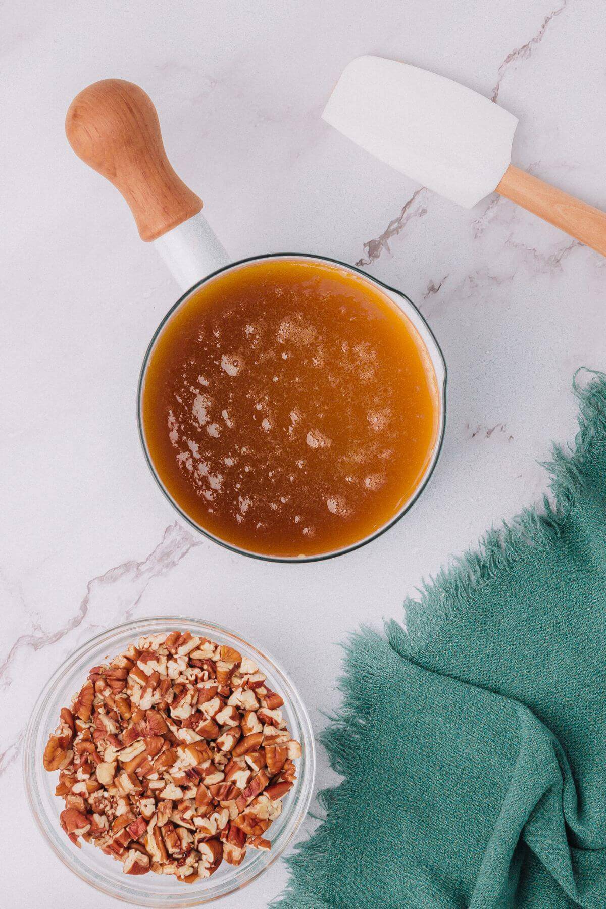 A saucepan with caramel sauce next to a bowl of chopped pecans and a spatula on a marble surface.