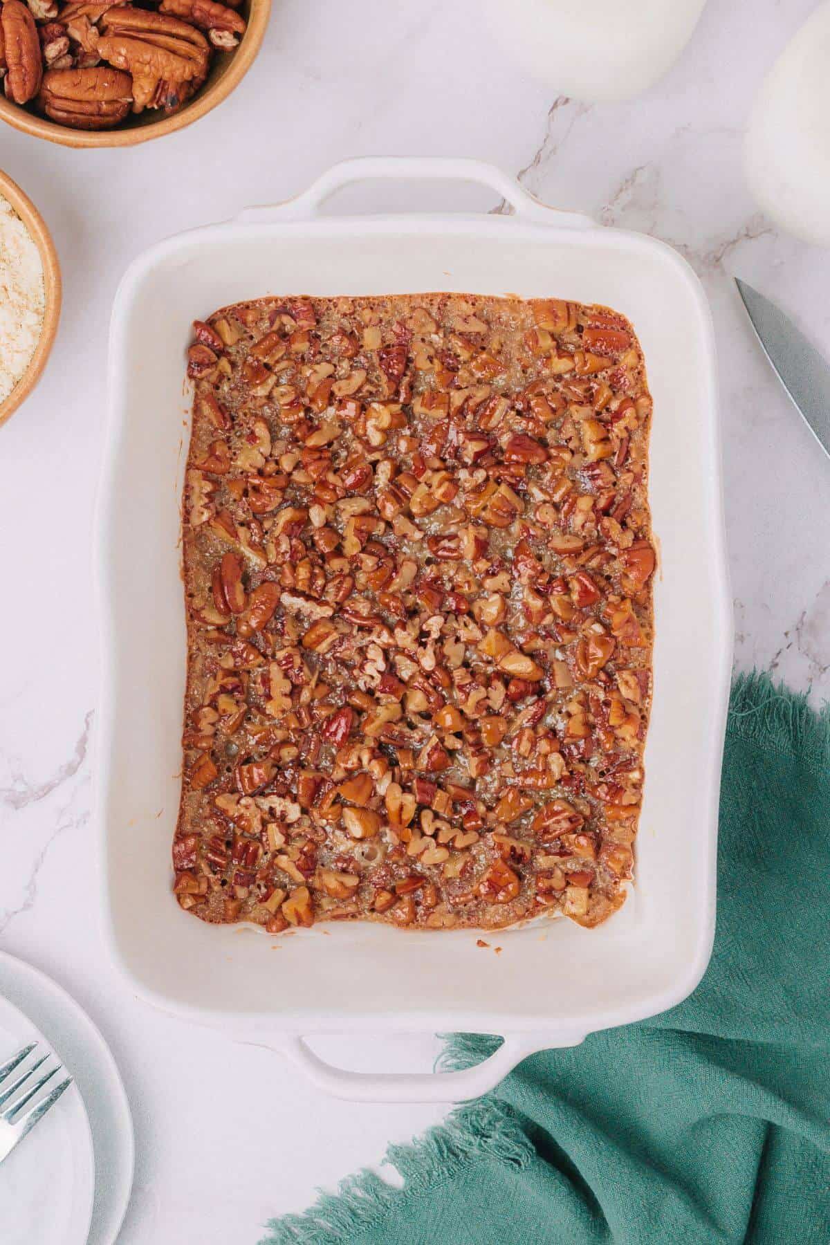 A rectangular baking dish contains pecan pie bars with a golden crust and a topping of pecans. A green cloth and utensils are partially visible beside it.