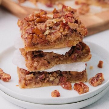 A stack of pecan bars on a white plate, with chopped pecans on top and layers of paper between the bars.