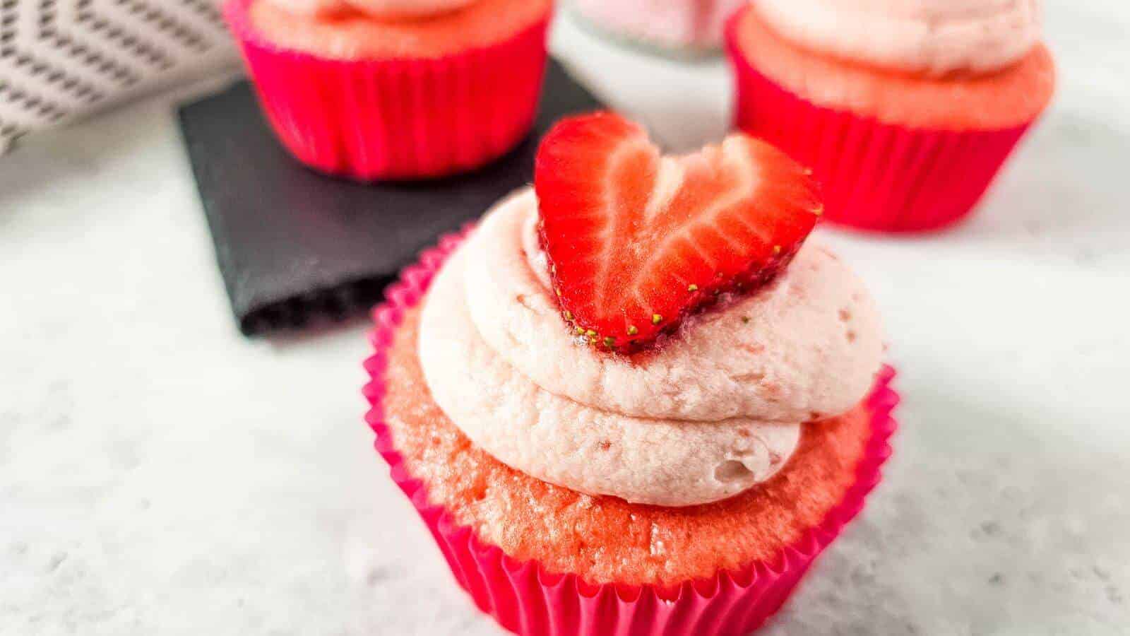 A pink cupcake with strawberry frosting topped with a halved strawberry in a pink wrapper, placed on a white surface. Two more cupcakes are blurred in the background.