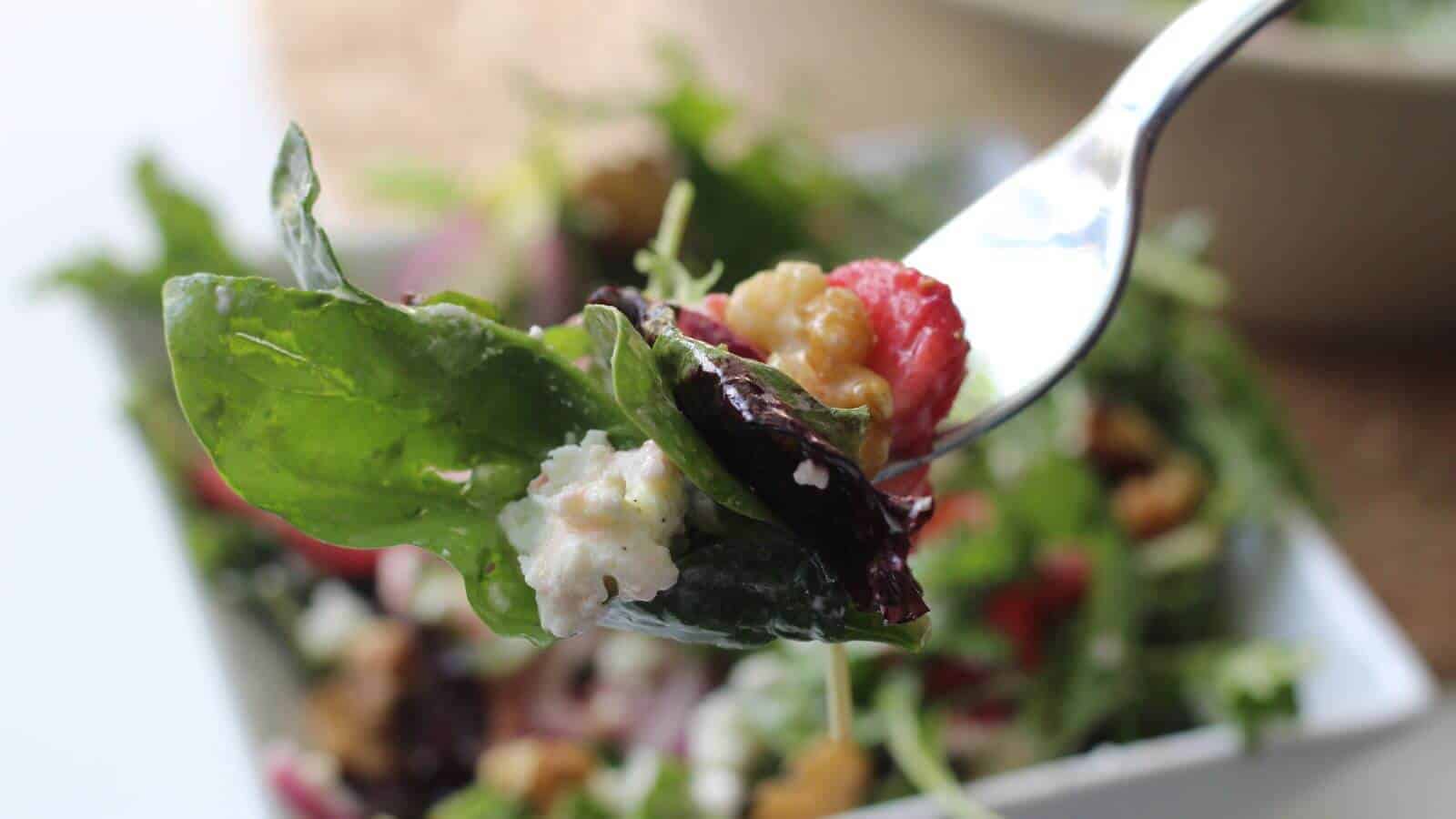 Close-up of a fork holding salad with greens, feta cheese, strawberries, and walnuts, with a bowl of salad blurred in the background.