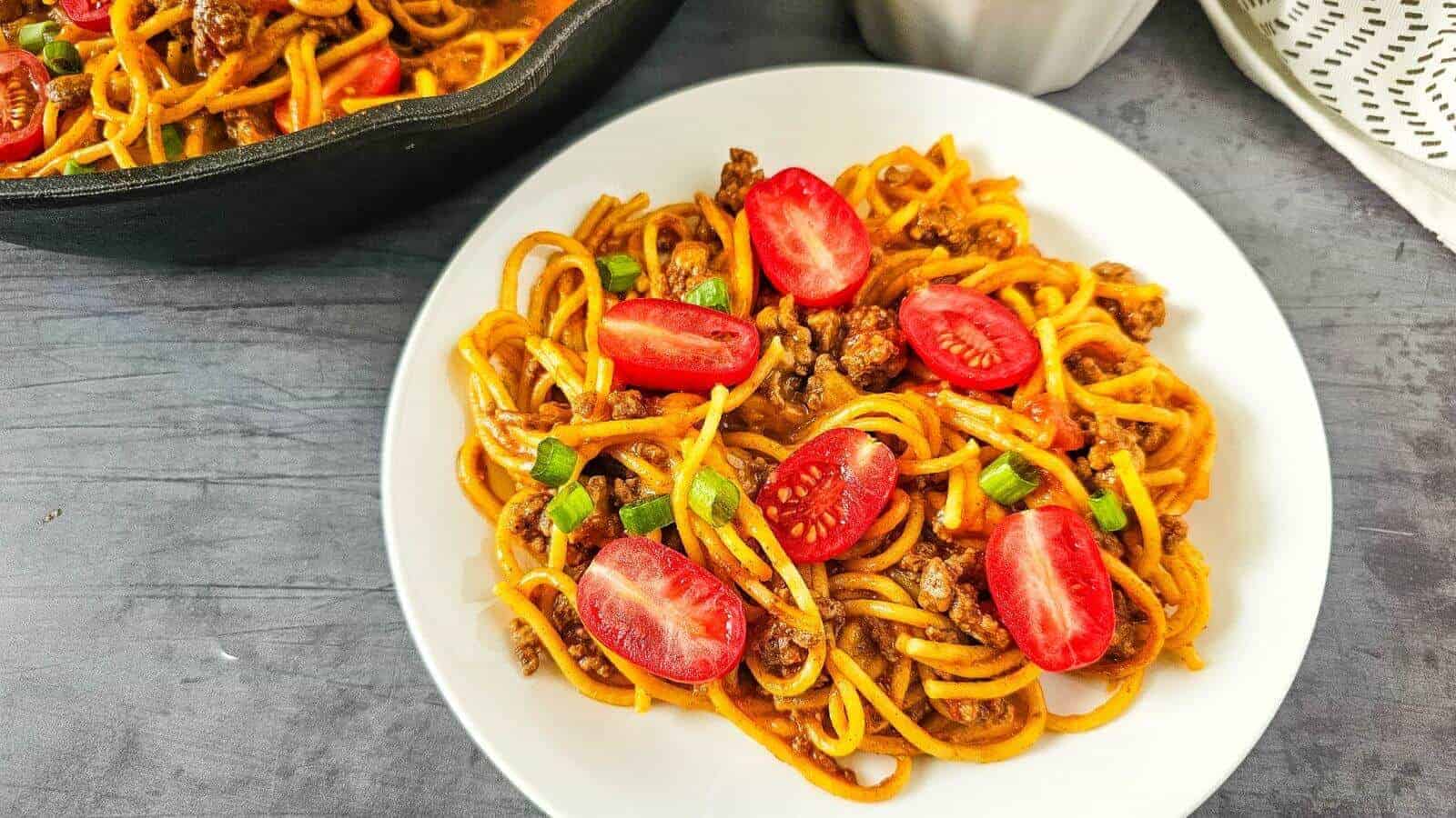 A plate of spaghetti with ground meat, halved cherry tomatoes, and chopped green onions, served next to a black skillet and a white napkin on a gray surface.
