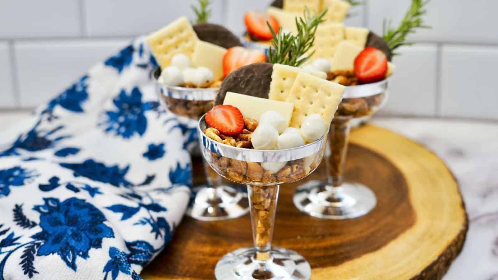 Three goblets filled with crackers, nuts, cheese, chocolate, strawberries, and rosemary garnish, placed on a wooden board with a blue floral cloth nearby.