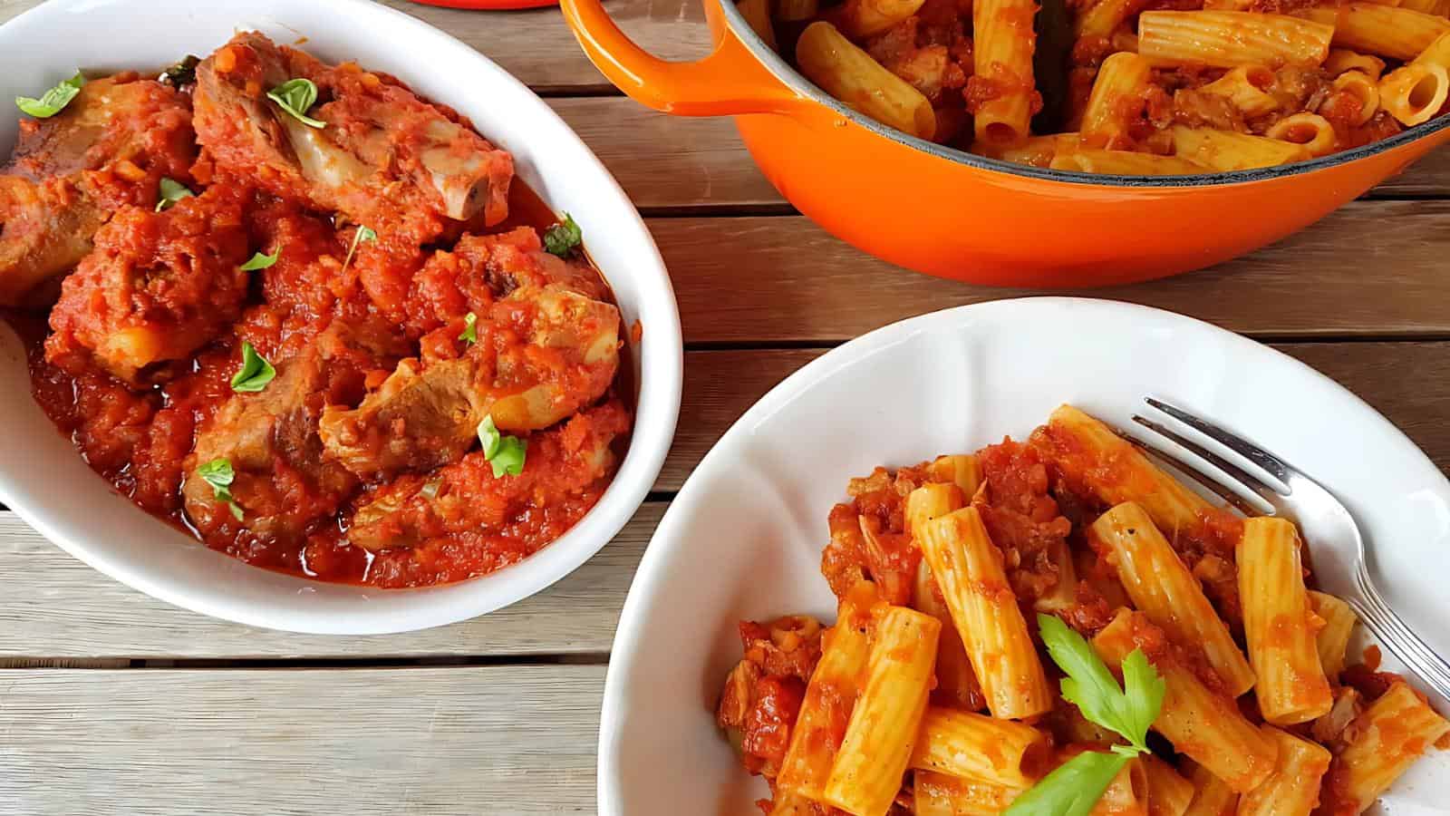 Cooked rigatoni pasta in tomato sauce is served on a white plate. A pot and a dish of meat in tomato sauce are placed nearby on a wooden table.