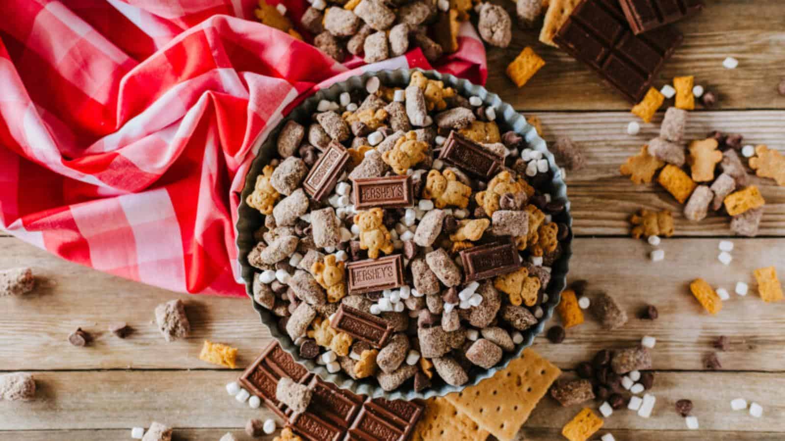 Bowl of snack mix with chocolate pieces, cereal bits, and marshmallows on a wooden surface, surrounded by a red checkered cloth and scattered ingredients.