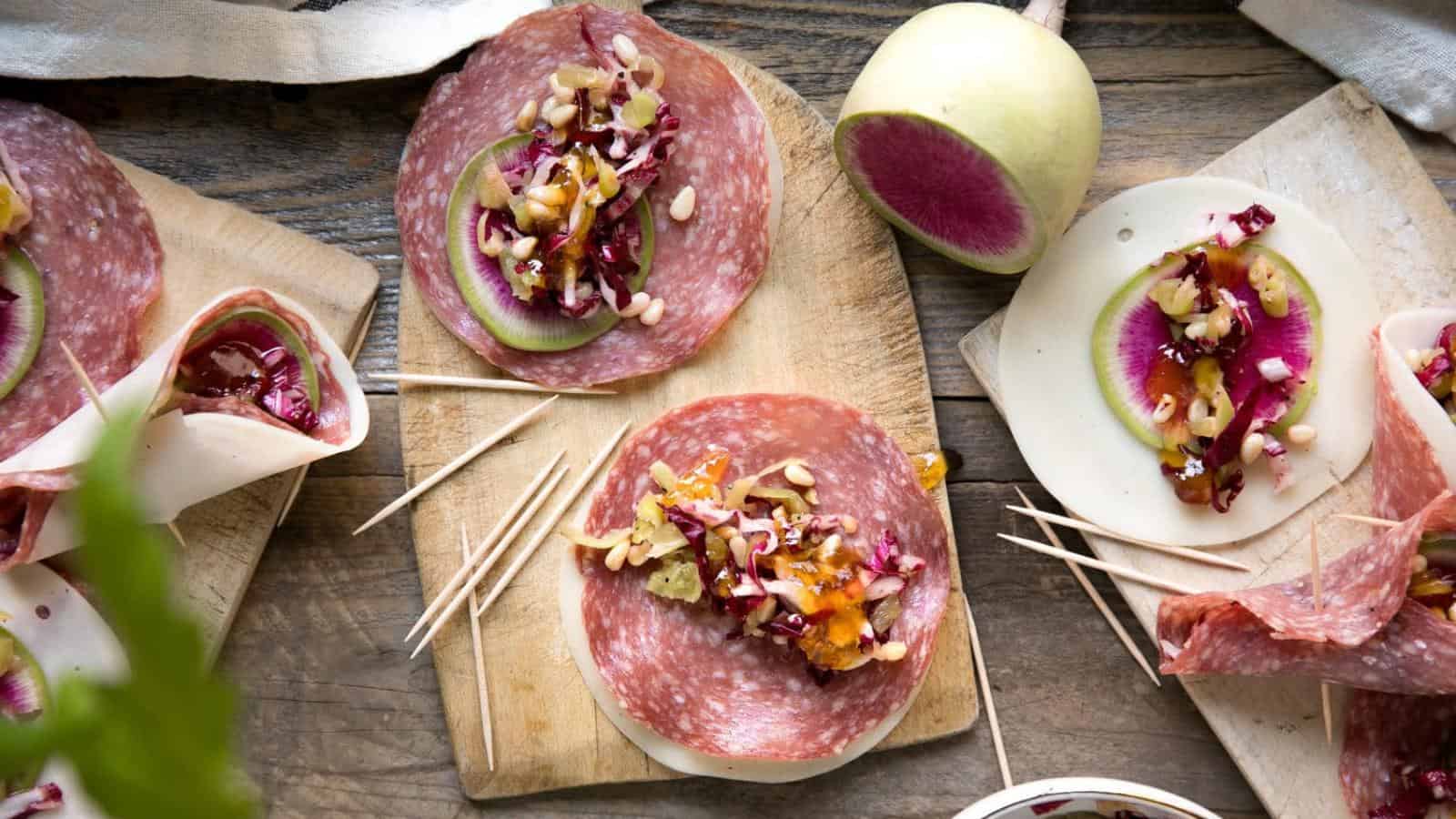 A wooden board with salami and cheese wraps filled with assorted vegetables, nuts, and seeds. A sliced radish and toothpicks are also present.