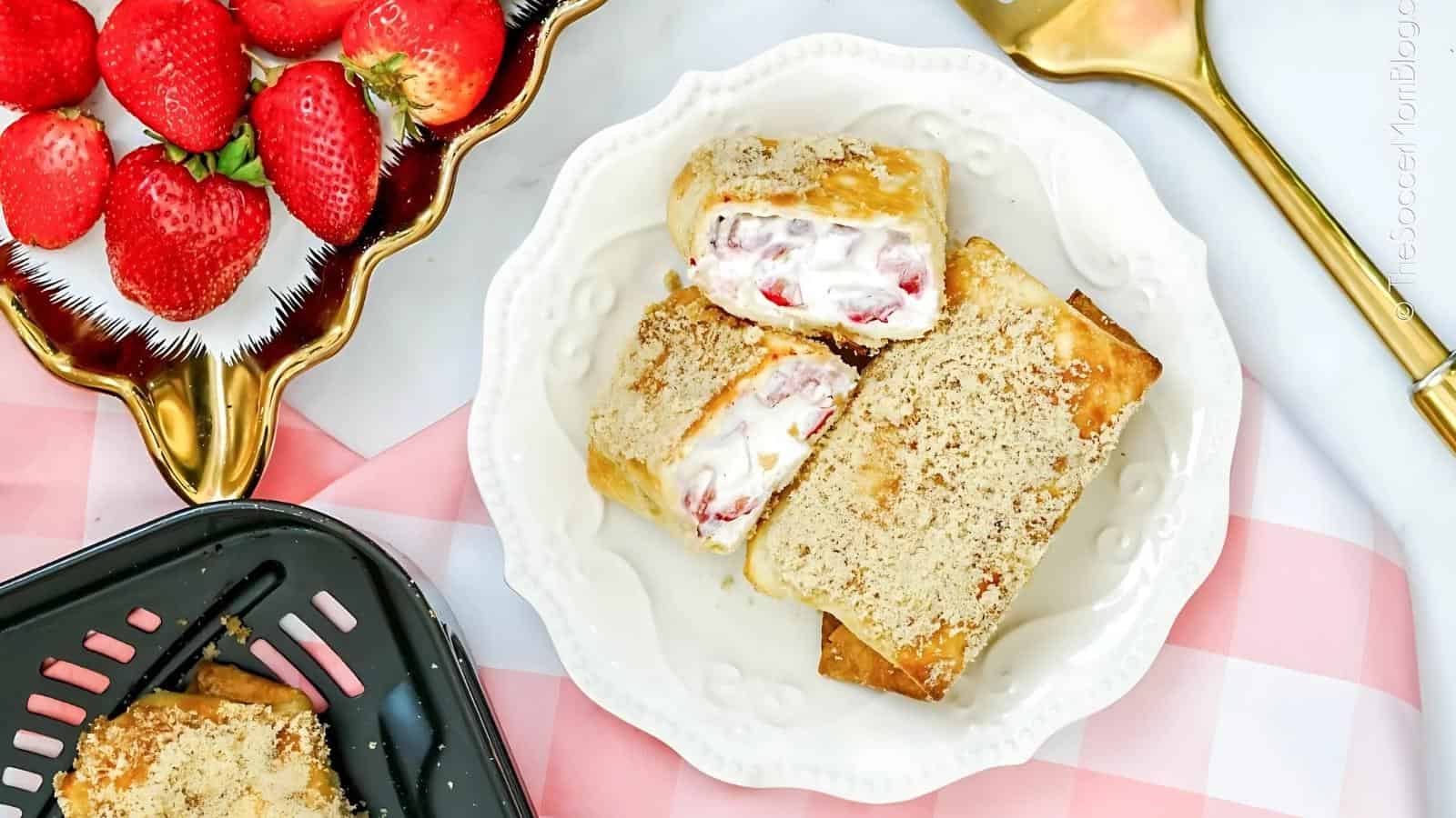 Two strawberry cream dessert chimichangas on a white plate next to a bowl of fresh strawberries and a baking dish on a pink checked cloth.