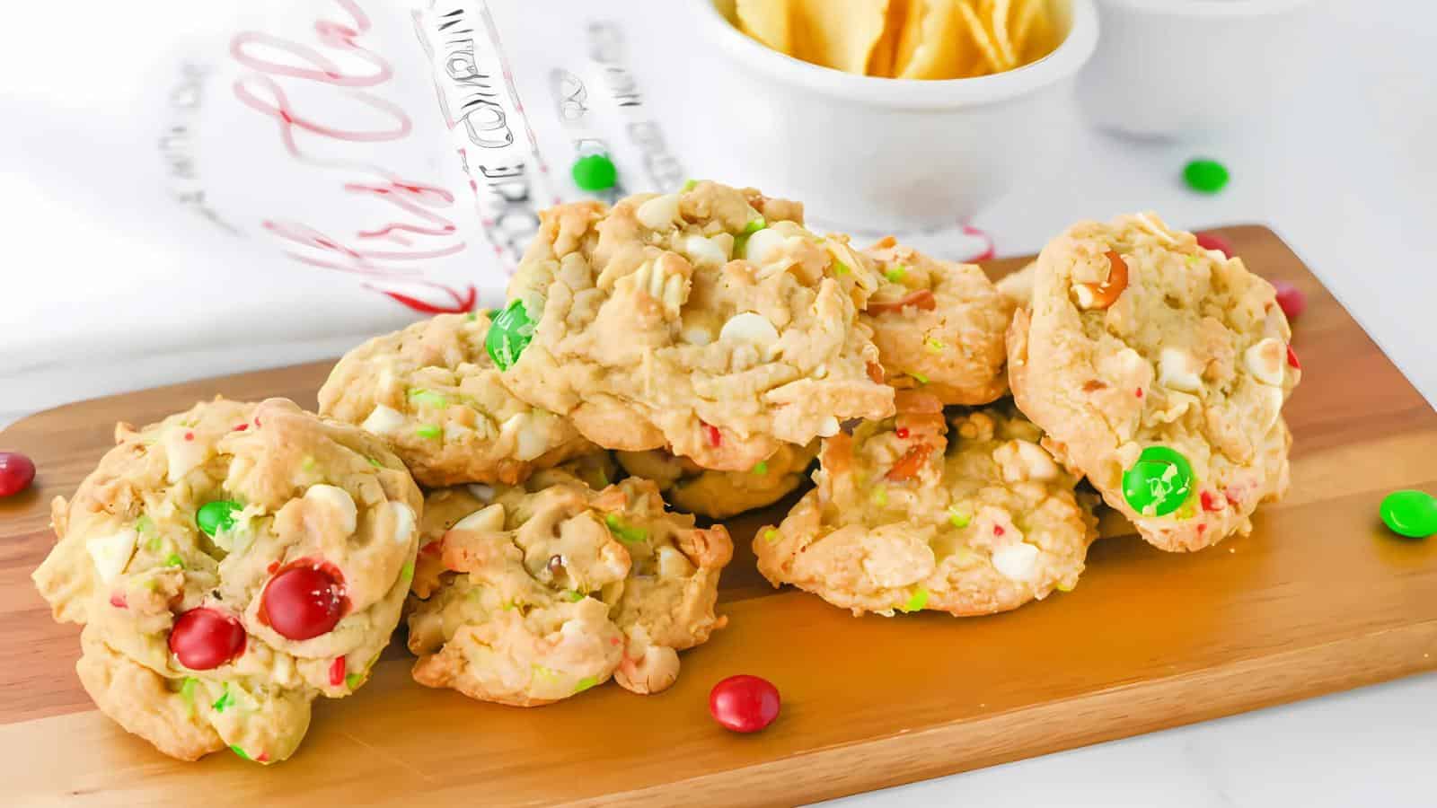 A stack of colorful candy cookies with white chocolate chips on a wooden board, accompanied by a white bowl in the background.