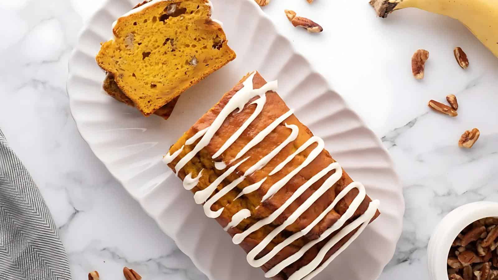 Loaf of pumpkin bread with white icing on a platter, garnished with pecans. Two slices are cut from the loaf.