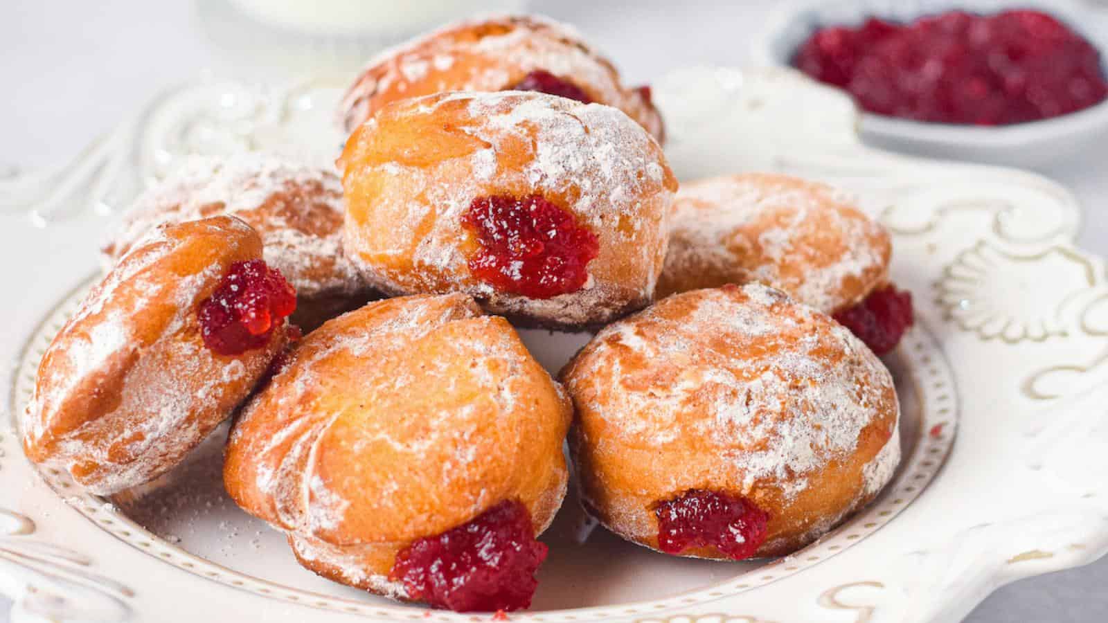 A plate of powdered jelly-filled donuts with a bowl of red jelly in the background.