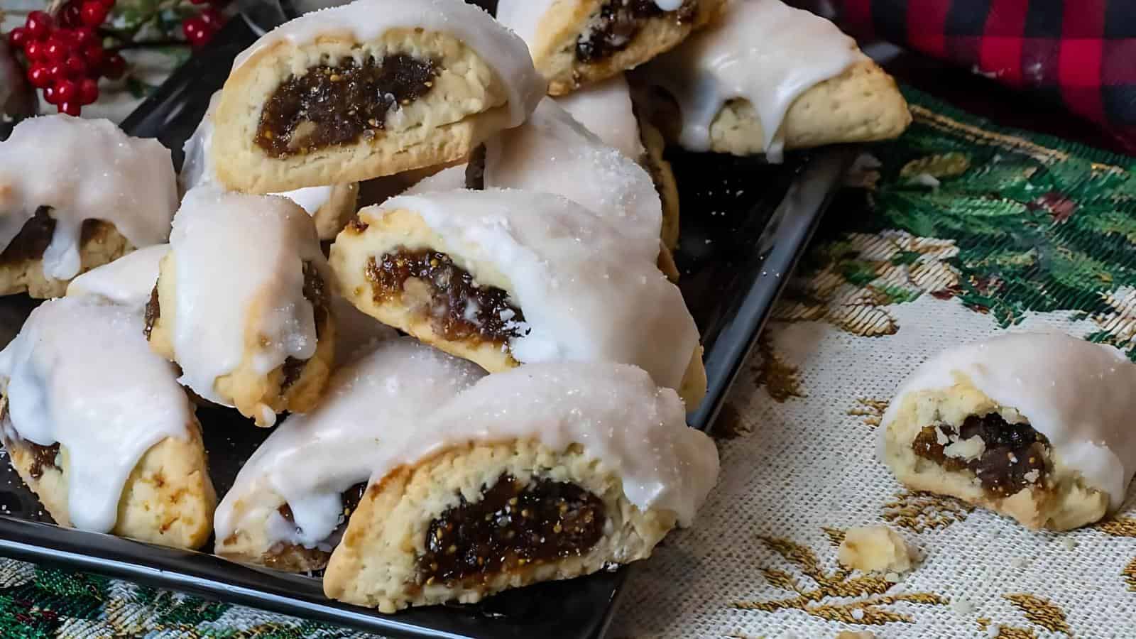 A tray of fig cookies with white icing is displayed on a tablecloth adorned with a green and gold pattern. One cookie is placed separately on the tablecloth.