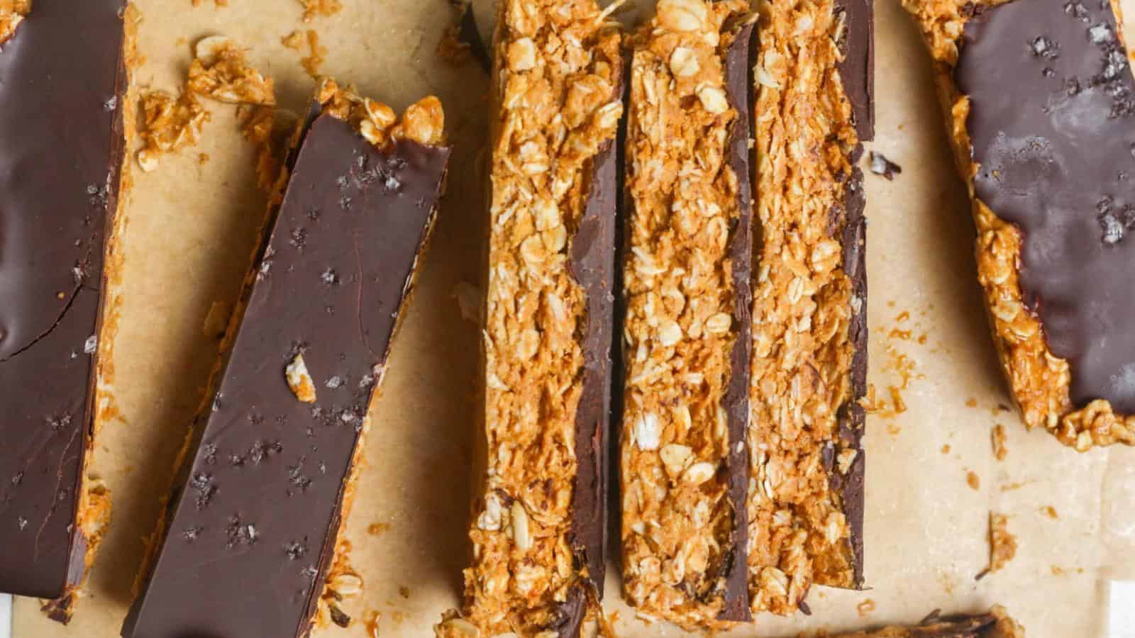 Granola bars with dark chocolate coating arranged on a parchment surface. Some are cut into slices, showing a crunchy oat texture.