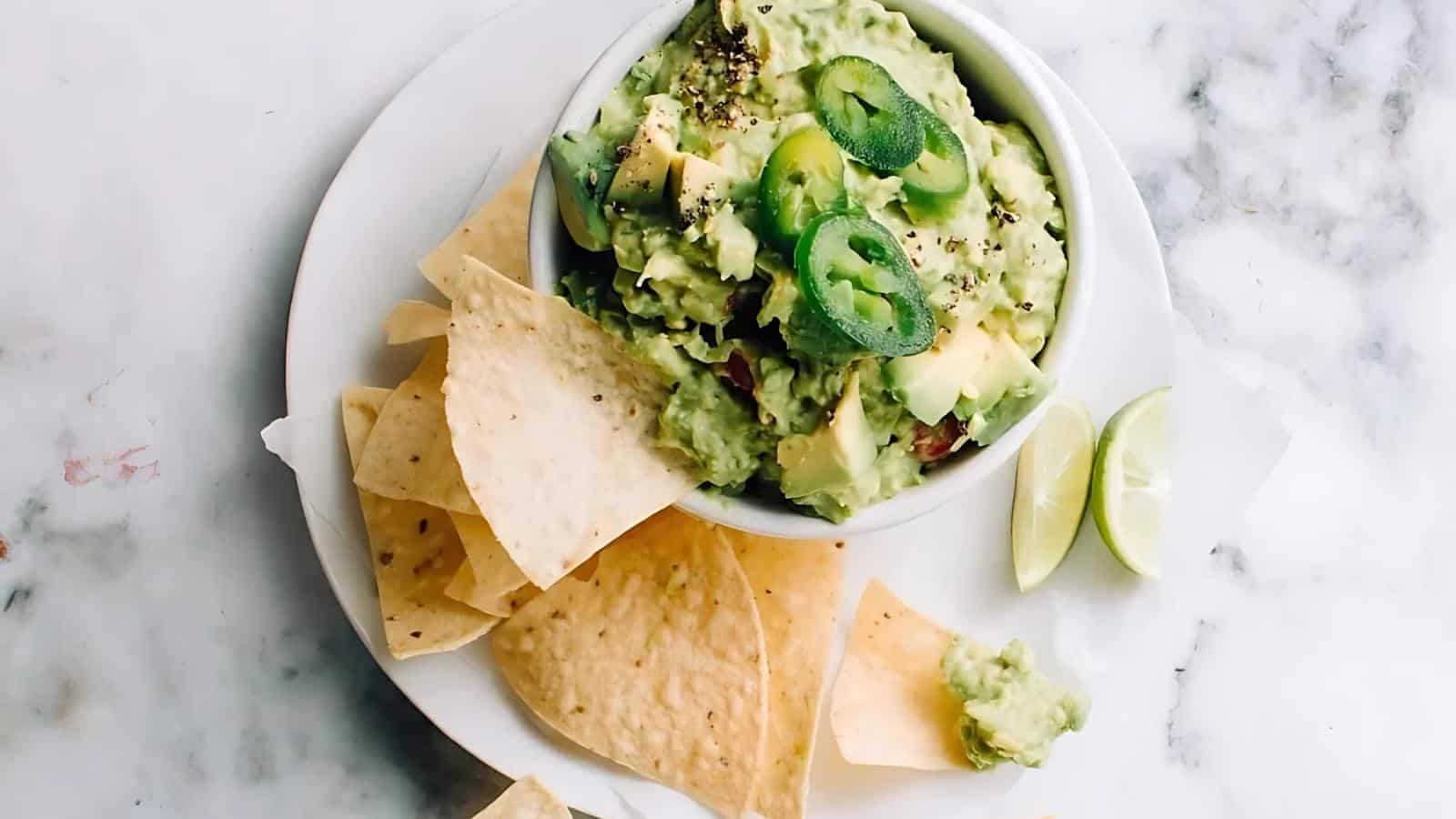 A bowl of guacamole topped with jalapeño slices is surrounded by tortilla chips and lime wedges on a white plate.