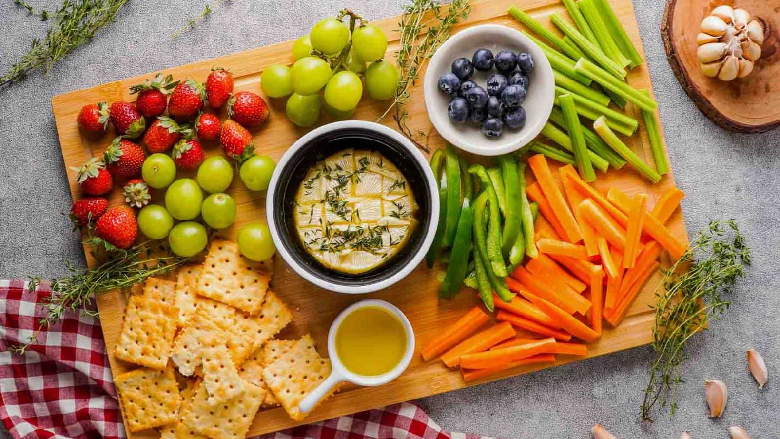 A wooden board with crackers, grapes, strawberries, blueberries, celery, green peppers, carrot sticks, and a bowl of cheese. A small jug of olive oil is nearby, and herbs are scattered around.