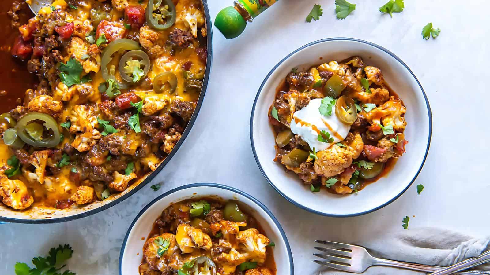 Bowl of baked cauliflower with beef, cheese, and jalapeños next to a skillet with more of the dish. Garnished with sour cream and cilantro. Silverware and lime nearby.