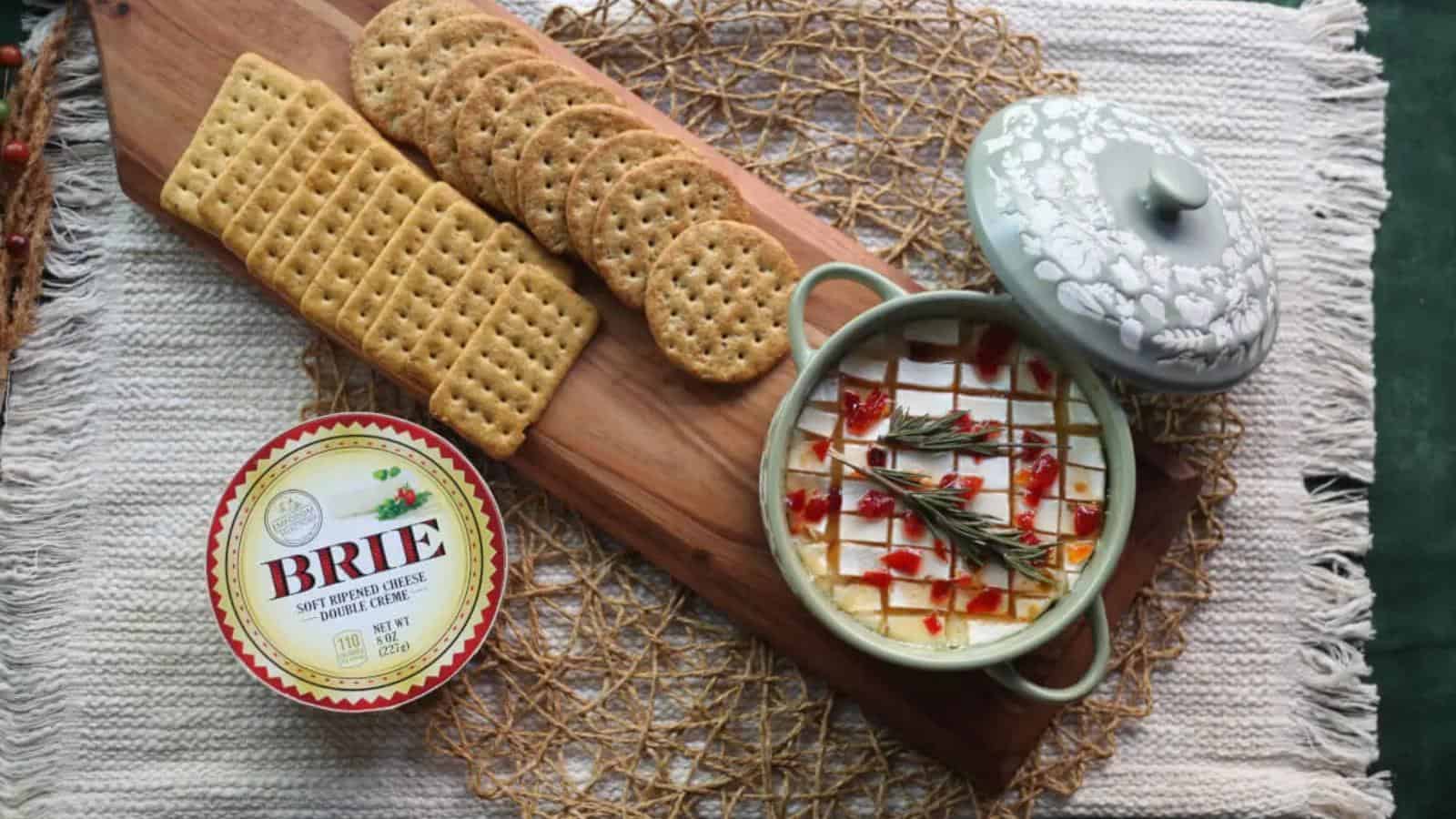 Cheese wheel with herbs and red garnish in a ceramic dish, crackers on a wooden board, and a Brie container on a woven mat.