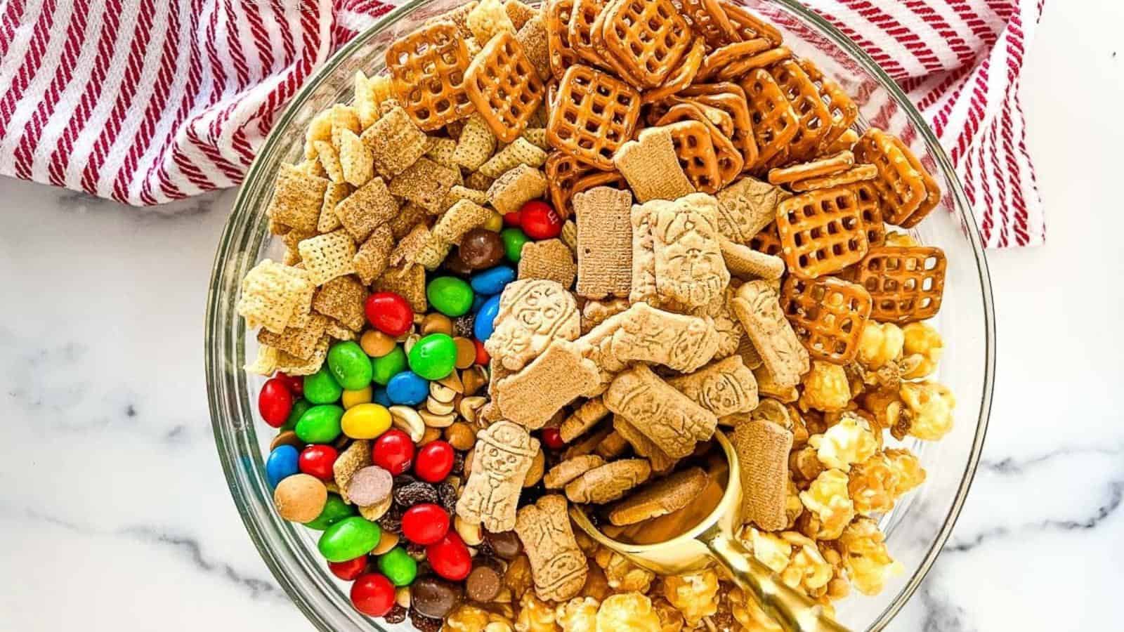 A bowl of mixed snacks including candy-coated chocolates, popcorn, pretzels, cereal squares, and animal-shaped crackers, with a gold scoop in the center.