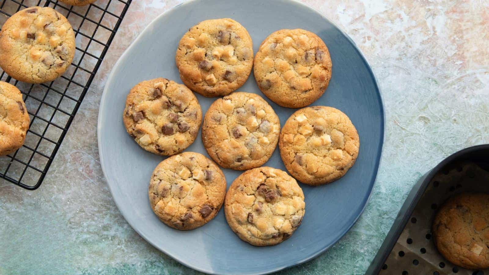 Seven chocolate chip cookies arranged on a round blue plate, surrounded by a baking tray and cooling rack on a textured surface.