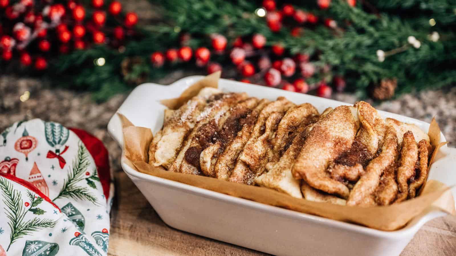 A ceramic dish of cinnamon pull-apart bread sits on a table near holiday-themed oven mitts, with festive decorations and greenery in the background.