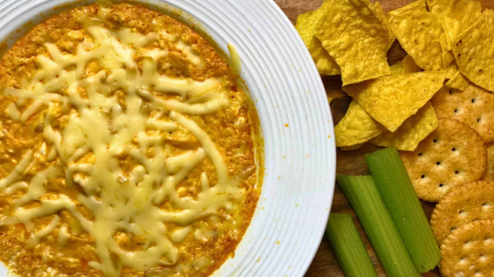 A plate of cheese dip with shredded cheese on top, served with tortilla chips, crackers, and celery sticks on the side.
