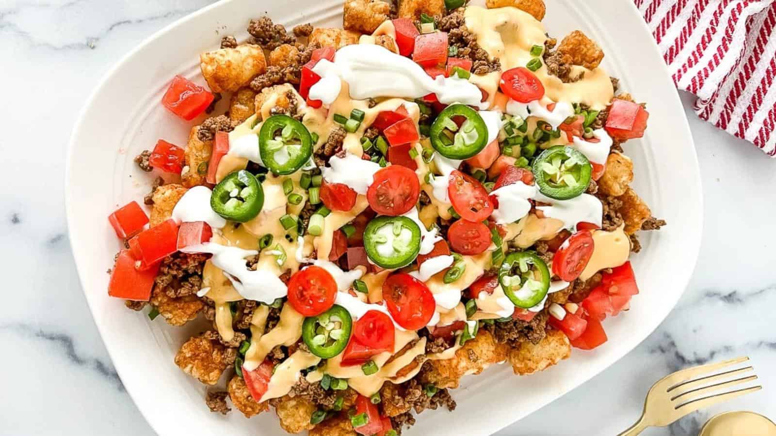 A plate of loaded nachos topped with ground beef, cheese sauce, sour cream, sliced jalapeños, cherry tomatoes, and green onions.