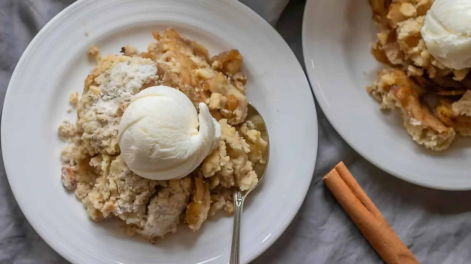 Two plates of apple crumble topped with vanilla ice cream on a table. A cinnamon stick is placed beside one of the plates.