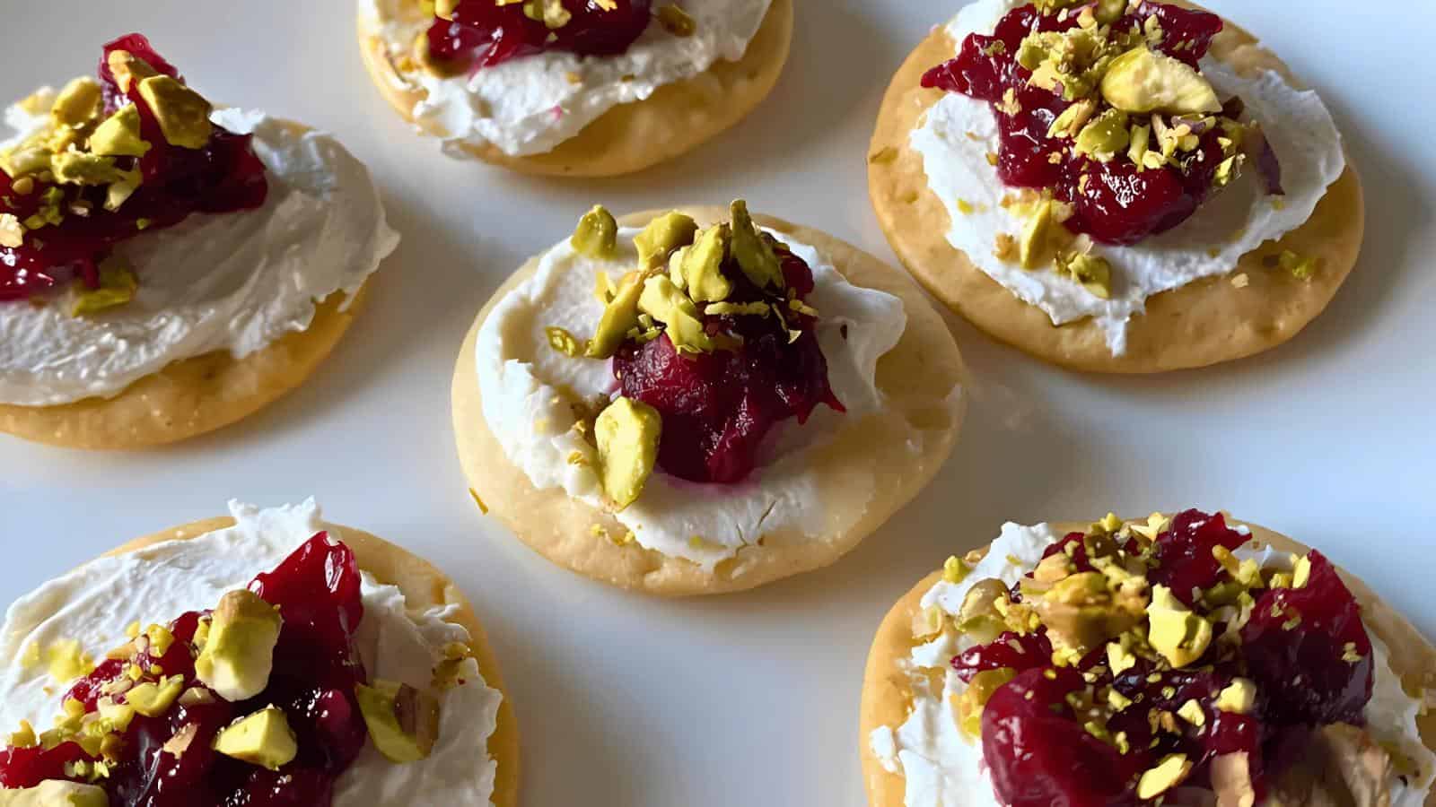 Round crackers topped with cream cheese, cranberry sauce, and chopped pistachios are arranged on a white plate.