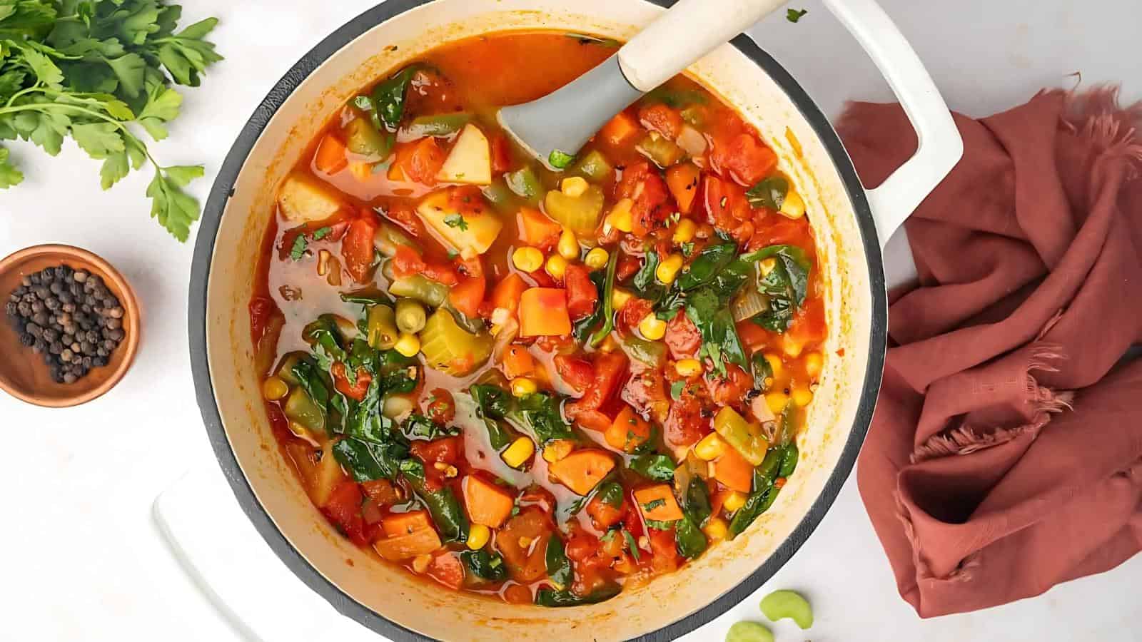 A pot of vegetable soup with spinach, carrots, corn, peas, and tomatoes on a white surface. Nearby are parsley, black pepper, and a brown cloth.
