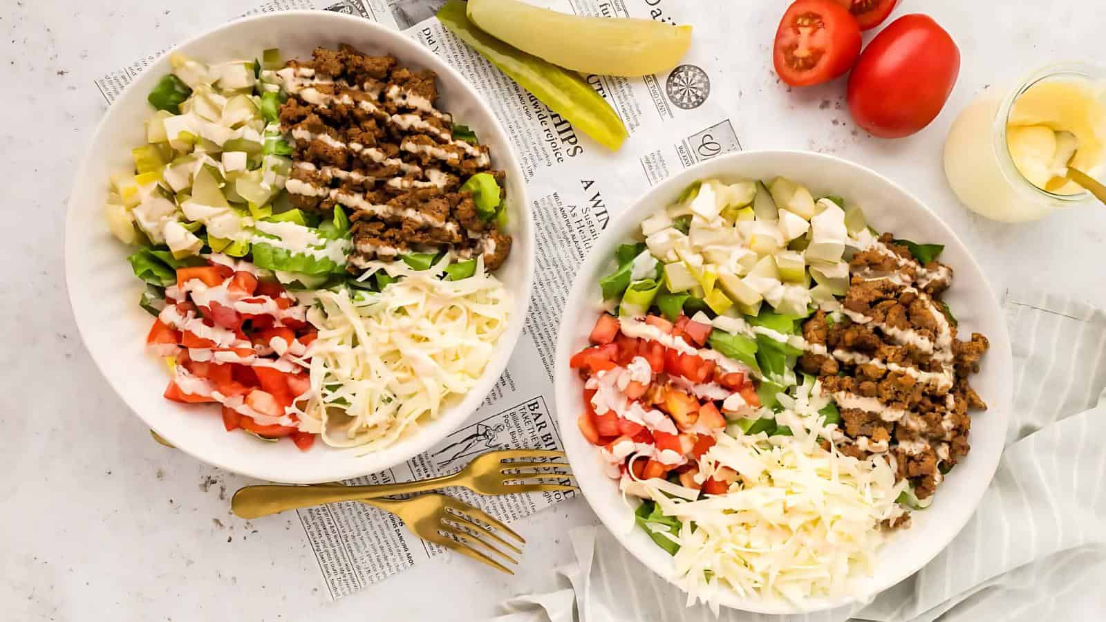 Two bowls of salad with ground meat, diced tomatoes, lettuce, avocado, cheese, and drizzled dressing. Forks, a pickle, tomatoes, and a dressing container are nearby.
