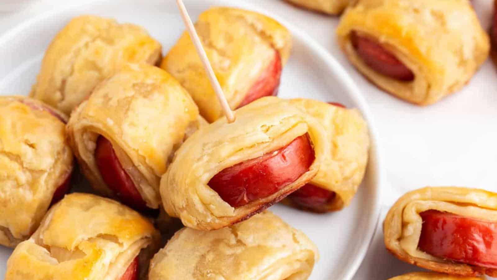 A plate of pigs in a blanket featuring small sausages wrapped in golden brown pastry, with one piece skewered by a toothpick.