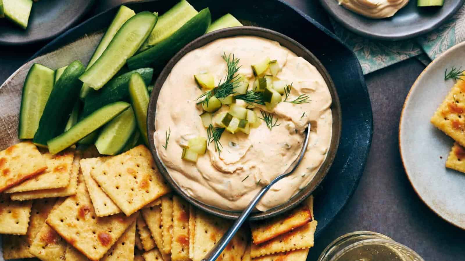 A bowl of creamy dip topped with diced pickles and dill, surrounded by cucumber slices and salted crackers on a platter.