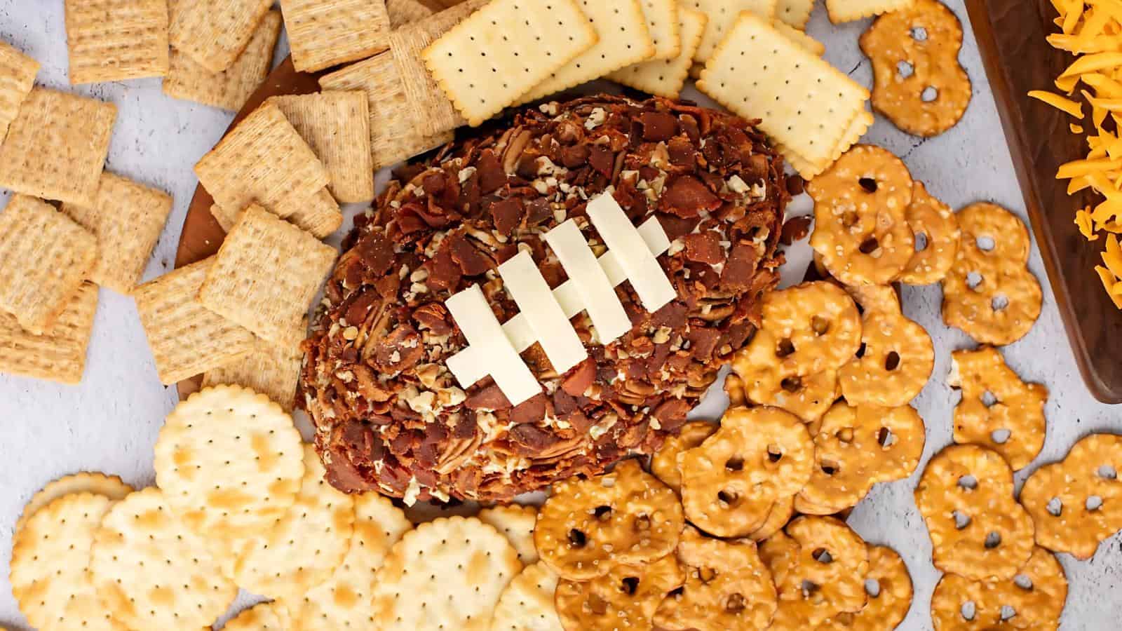 A cheese ball shaped like a football, coated with nuts and topped with cheese strips, surrounded by an assortment of crackers and pretzels.