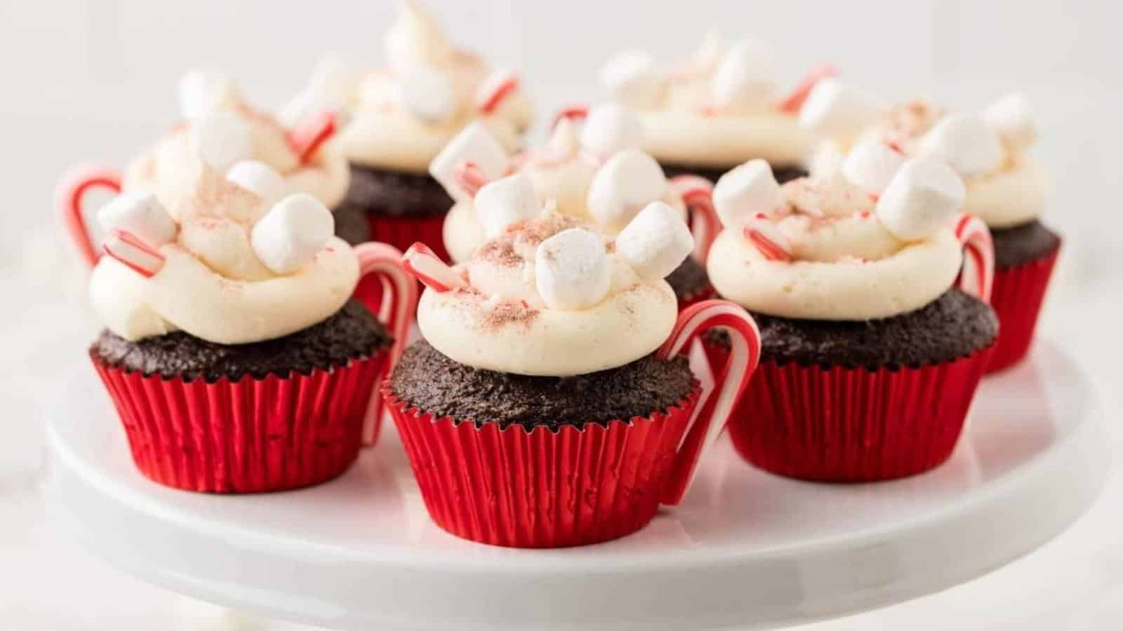 Chocolate cupcakes with white frosting, topped with mini marshmallows, cocoa powder, and candy cane pieces, displayed on a white platter.