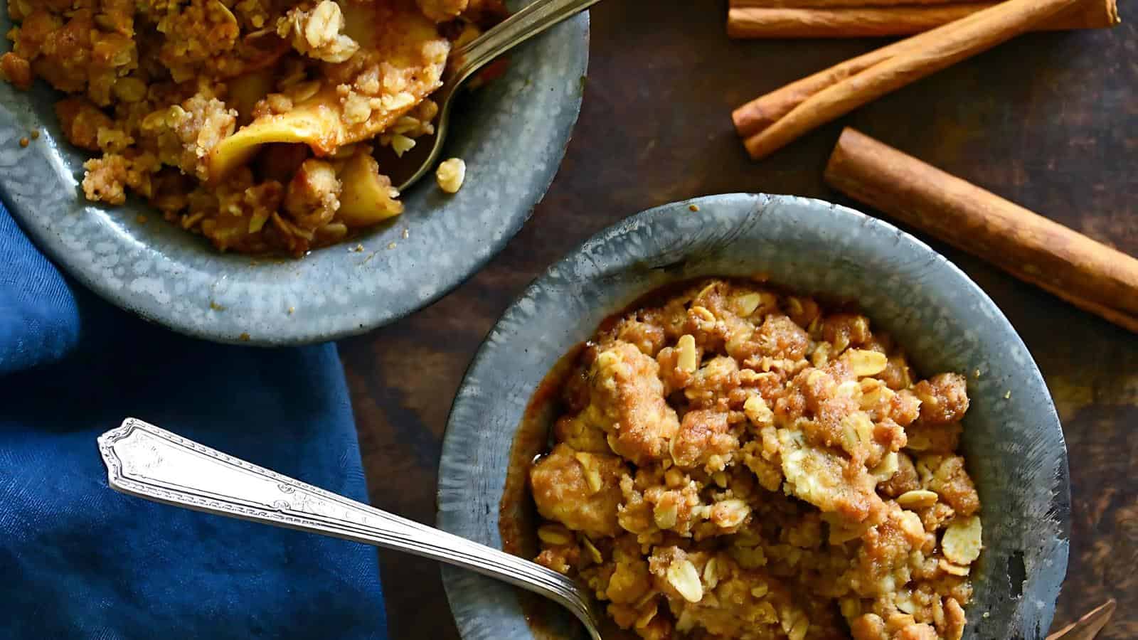 Two metal bowls of apple crumble with silver spoons sit on a dark surface, accompanied by cinnamon sticks and a blue cloth.