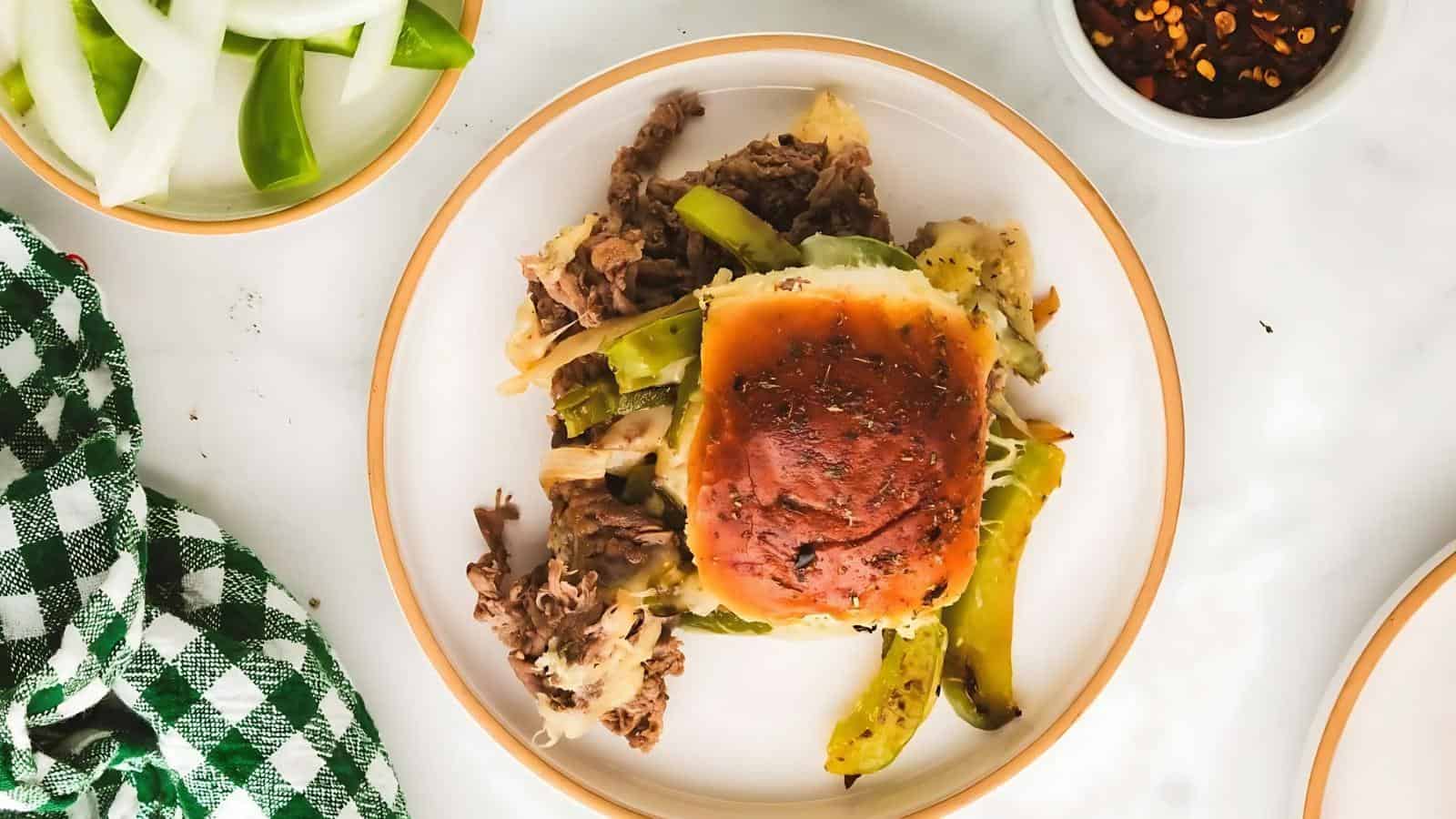 Plate with a sandwich on a bun, filled with shredded meat and green peppers, next to sliced onions and a bowl of red pepper flakes. Green checked cloth on the side.