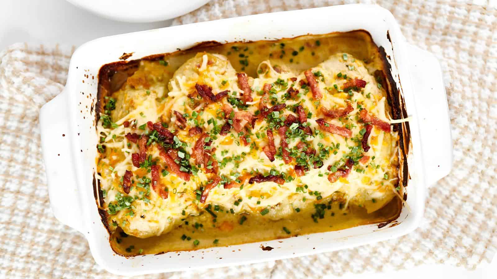 A baked dish with stuffed chicken breasts topped with melted cheese, bacon bits, and chopped herbs in a white baking dish on a beige woven cloth.