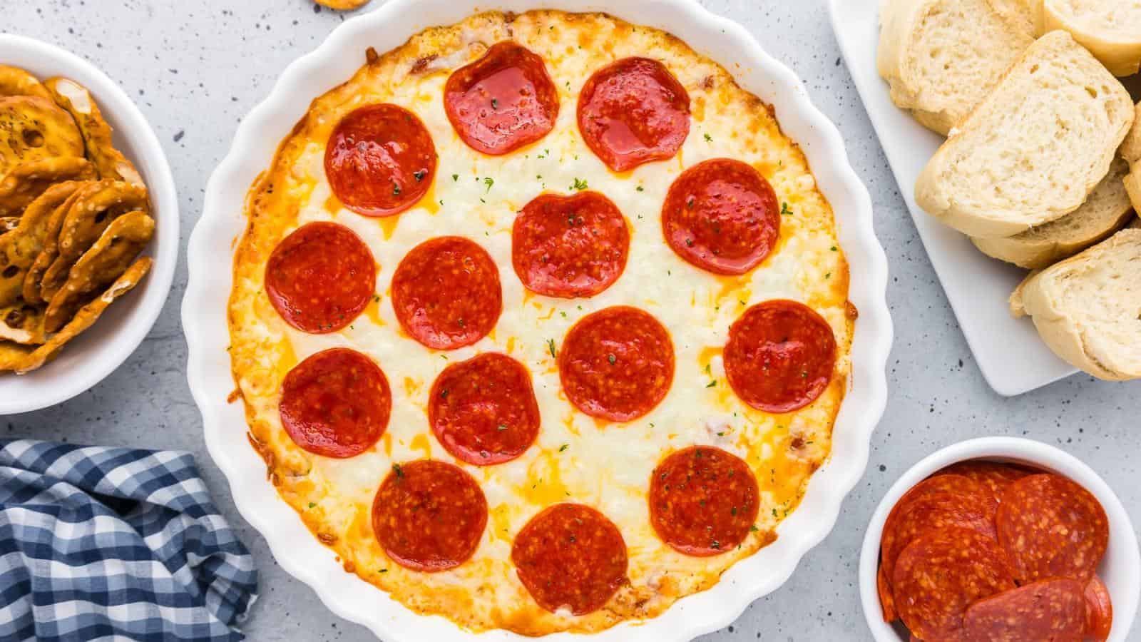 A pepperoni-covered dip in a round baking dish is surrounded by pretzels, sliced bread, and more pepperoni on a gray surface with a blue checkered cloth.