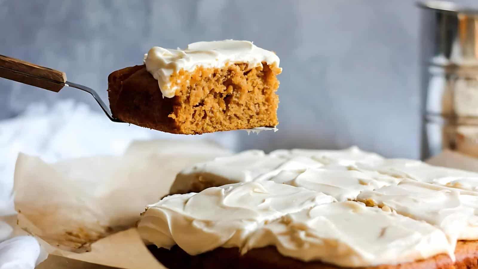 A slice of pumpkin cake with cream cheese frosting is lifted from a larger cake on parchment paper.