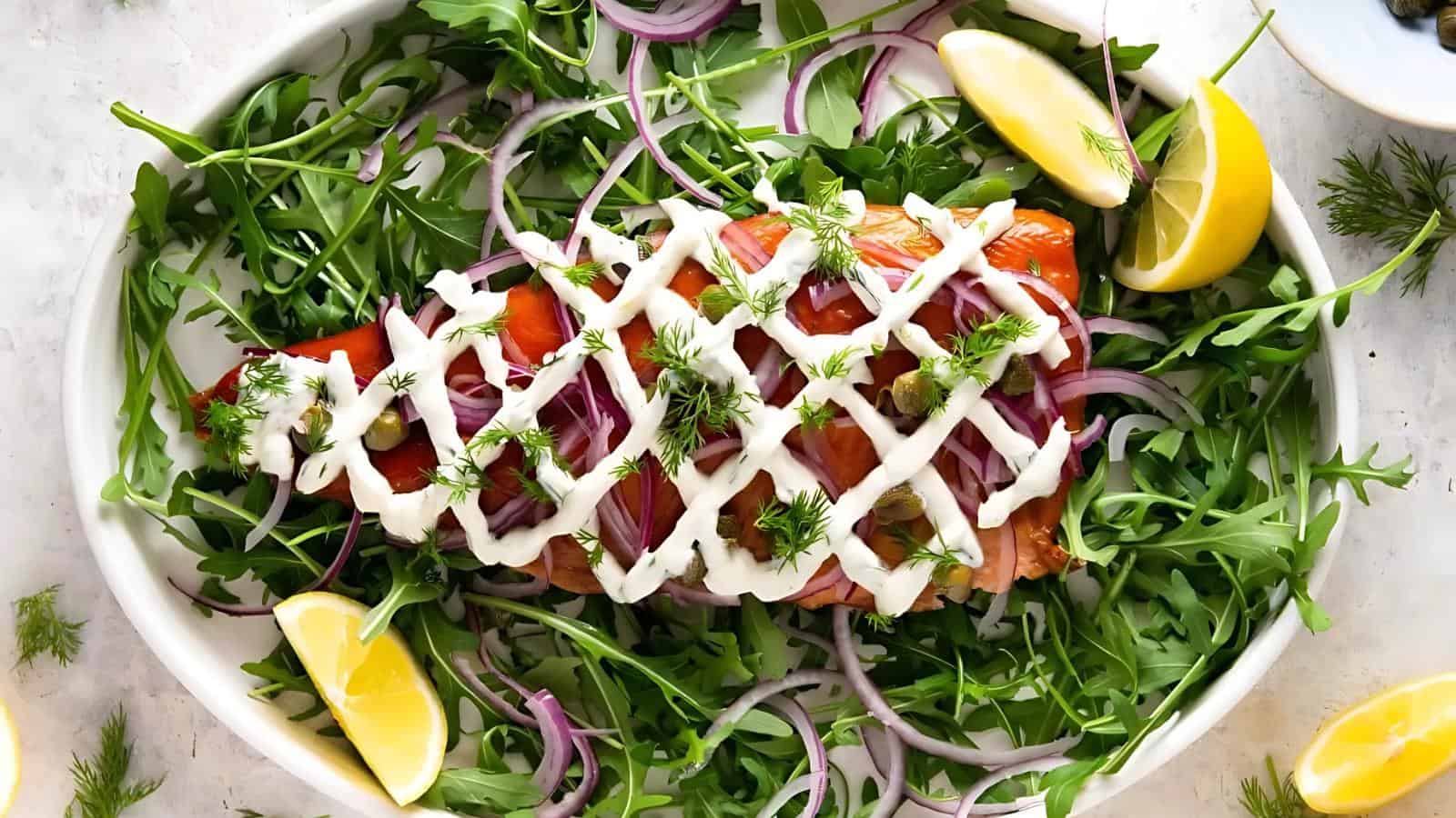 Salmon topped with creamy sauce, red onion slices, and dill on a bed of arugula with lemon wedges on a white plate.