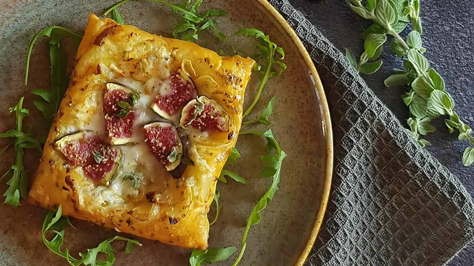 A square flatbread topped with figs and cheese on a beige plate, garnished with arugula, next to a textured cloth and sprigs of greens.
