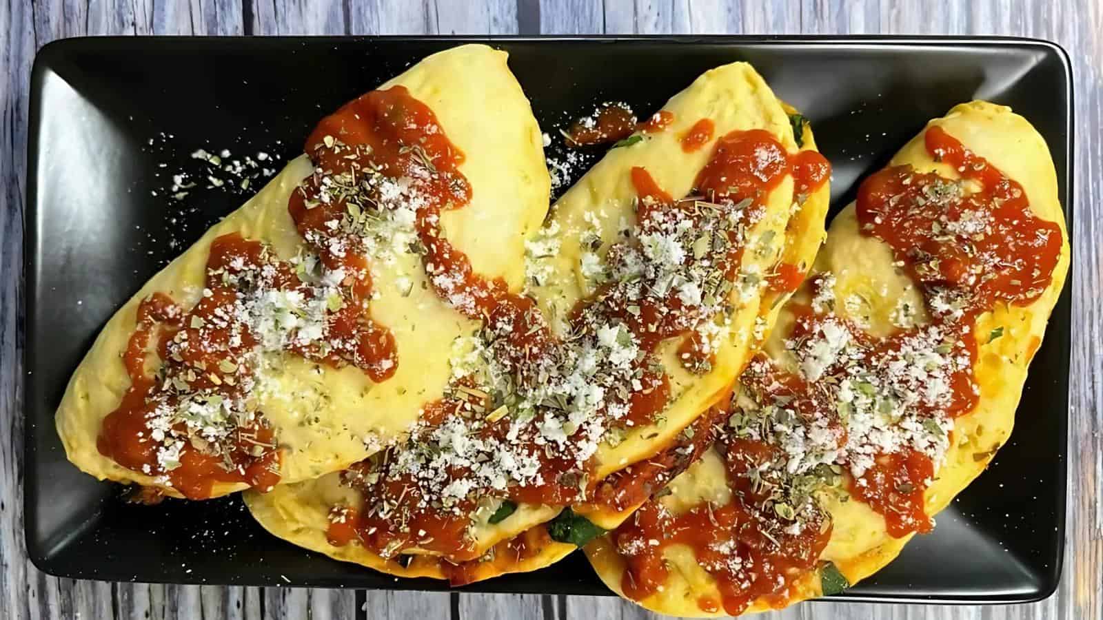 Three empanadas topped with tomato sauce and grated cheese on a black rectangular plate.